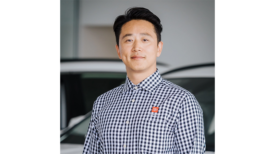 A person wearing a black-and-white checkered button-up shirt with a Toyota logo patch on the left chest. The background shows a blurred indoor setting with parts of white vehicles visible, suggesting a car showroom or dealership environment