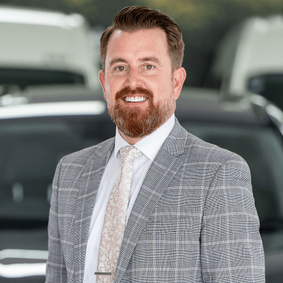 Person wearing a light gray checkered suit jacket, white dress shirt, and patterned light-colored tie, standing in front of blurred vehicles