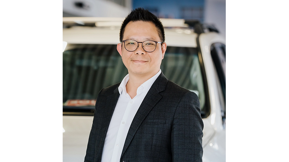Person wearing a dark checked blazer over a white collared shirt, standing indoors in front of a white vehicle in what appears to be a showroom or automotive service area.