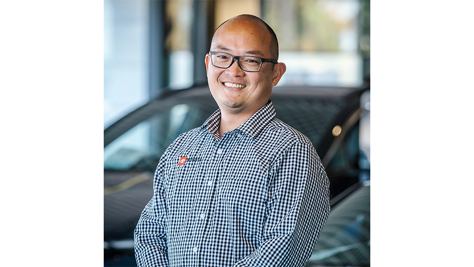 A person wearing a long-sleeve, black-and-white checkered button-up shirt with an embroidered logo on the left chest, standing indoors in front of a dark-colored car. The background shows a modern showroom environment with large windows and blurred outdoor scenery.