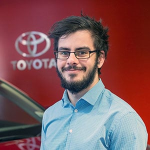 Toyota team member wearing a blue shirt and glasses, standing in front of a red Toyota sign.