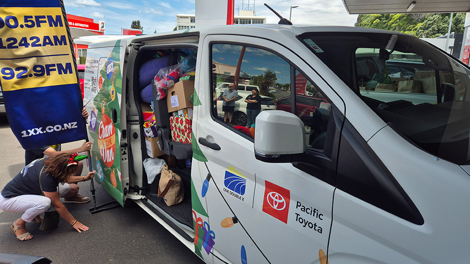 Pacific Toyota van packed with donated gifts and supplies during a community charity event, next to a banner showing local radio station frequencies and website.