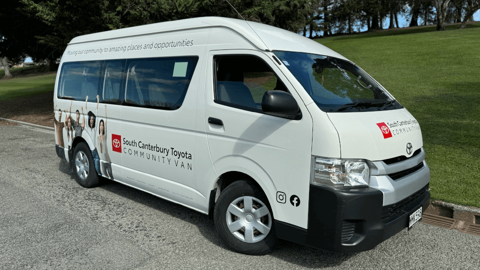 White Toyota community van parked on a paved surface outdoors, featuring branding for South Canterbury Toyota Community Van on the side. The van displays text reading ‘Moving our community to amazing places and opportunities’ along the top, along with social media icons for Instagram and Facebook near the rear. The design includes images of diverse people along the side panel. The background shows a grassy area with trees under daylight.
